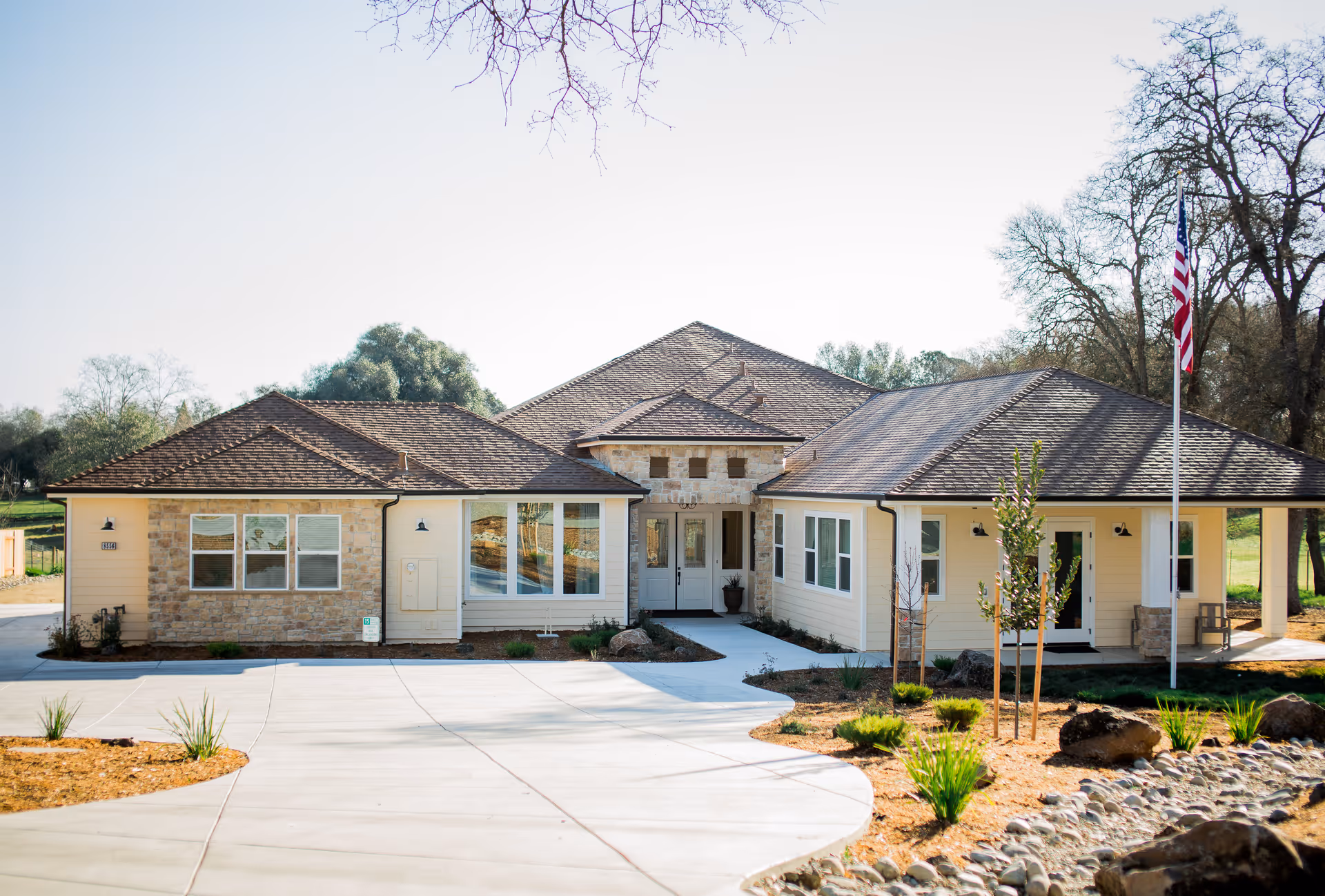 Front exterior view of a single-story building with a brown shingled roof, beige siding, and stone accents. The building has multiple windows and a central entrance with double doors. There is a concrete driveway and landscaped areas with small plants and rocks. An American flag is displayed on a flagpole to the right of the entrance.