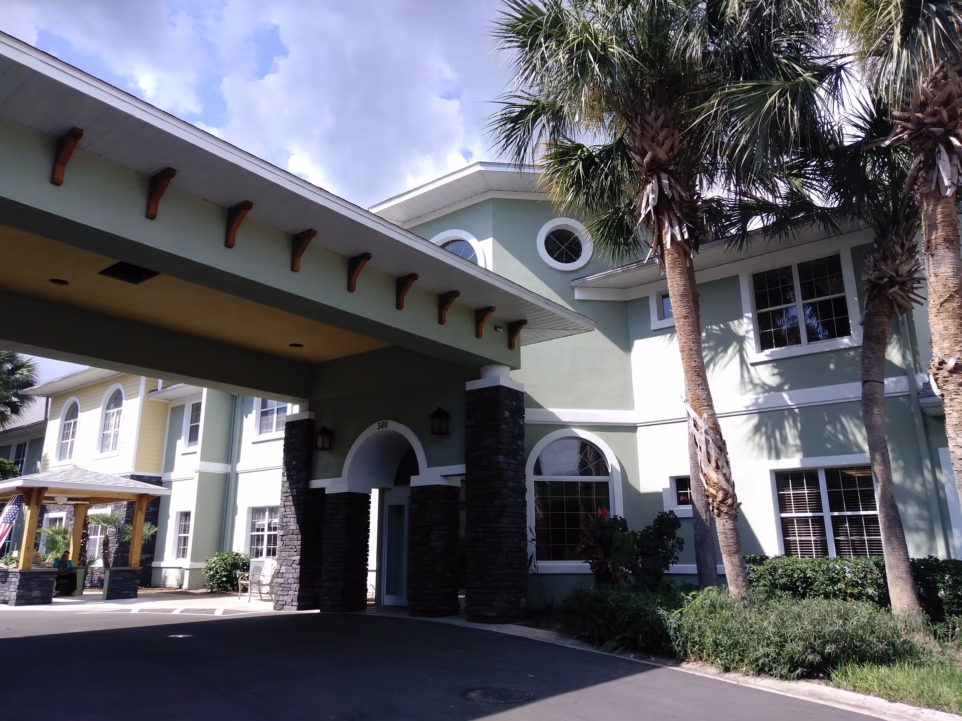 Exterior view of Bridgewater at Waterman Village Assisted Living facility showing a two-story building with light green walls, white trim, and stone accents around the entrance. There are palm trees and landscaping in front, and a covered driveway area under a roof supported by stone pillars. The sky is partly cloudy.