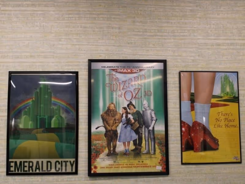 Three framed posters on a wall. The left poster shows an illustration of the Emerald City with a rainbow and the text 'EMERALD CITY'. The center poster is a promotional image for the 75th anniversary IMAX 3D showing of 'The Wizard of Oz' featuring the main characters Dorothy, the Cowardly Lion, the Scarecrow, and the Tin Man. The right poster shows a close-up of Dorothy's legs wearing white socks and red ruby slippers on a yellow brick road with the text 'There's No Place Like Home.'