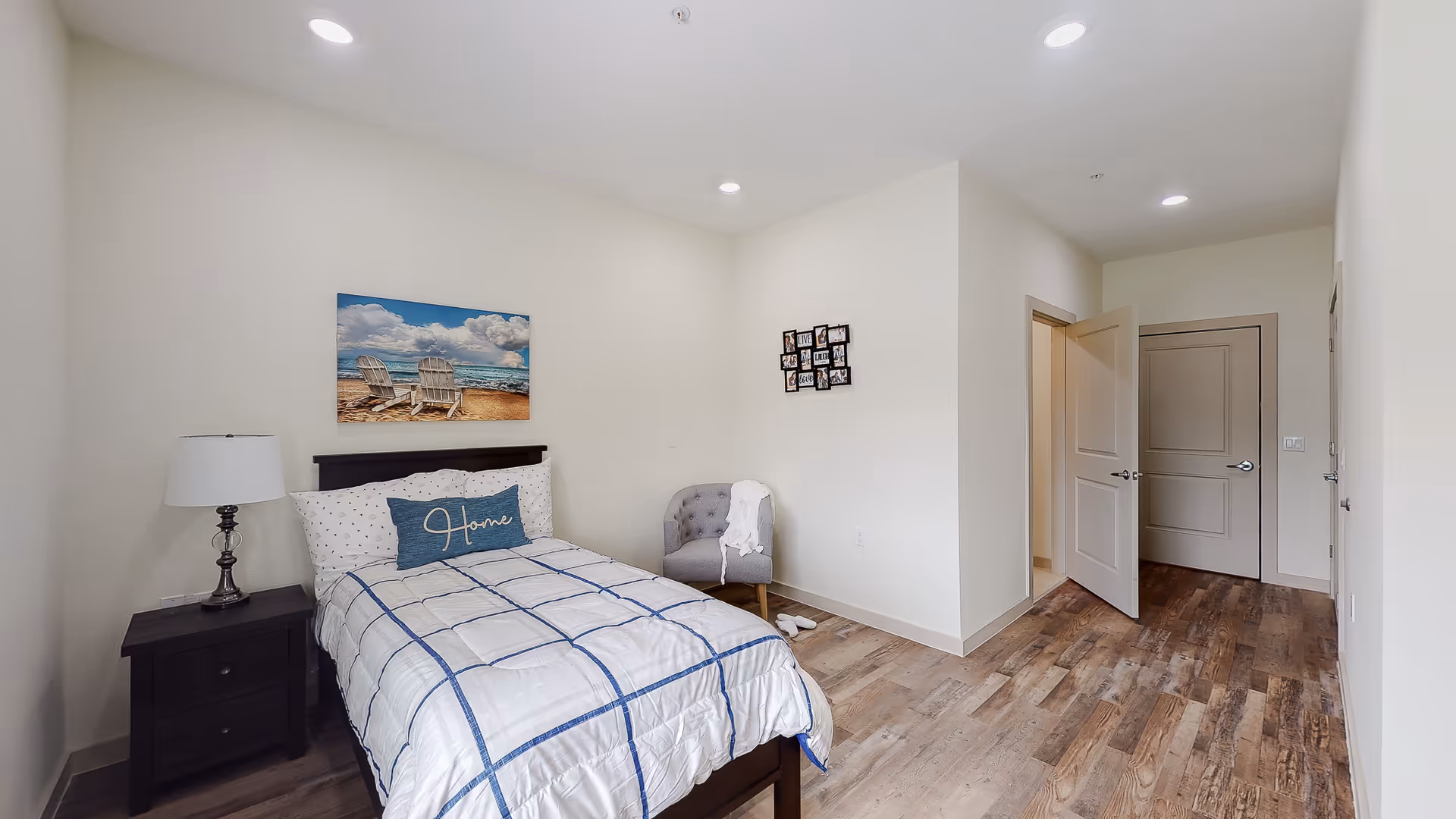 A neatly arranged bedroom with a bed featuring white and blue checkered bedding and a decorative pillow that says 'Home'. There is a dark wooden nightstand with a lamp on the left side of the bed. A gray upholstered chair with a white throw blanket is placed against the wall. On the wall above the bed is a beach-themed painting of two chairs facing the ocean. The room has light-colored walls, wood flooring, and two doors, one open and one closed.