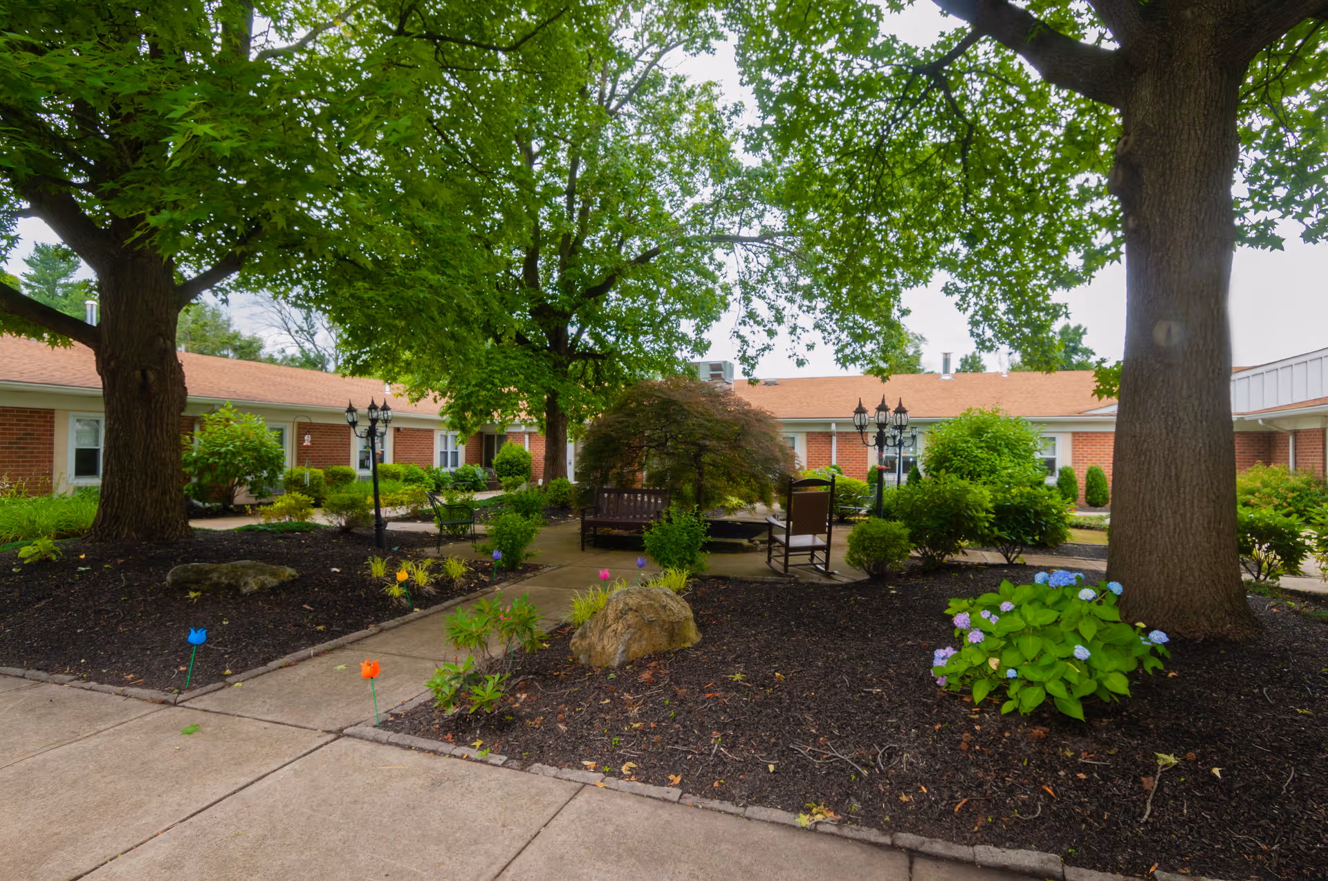 Shaded courtyard with large trees, benches, lamp posts, and landscaped flower beds in front of a single-story brick building.