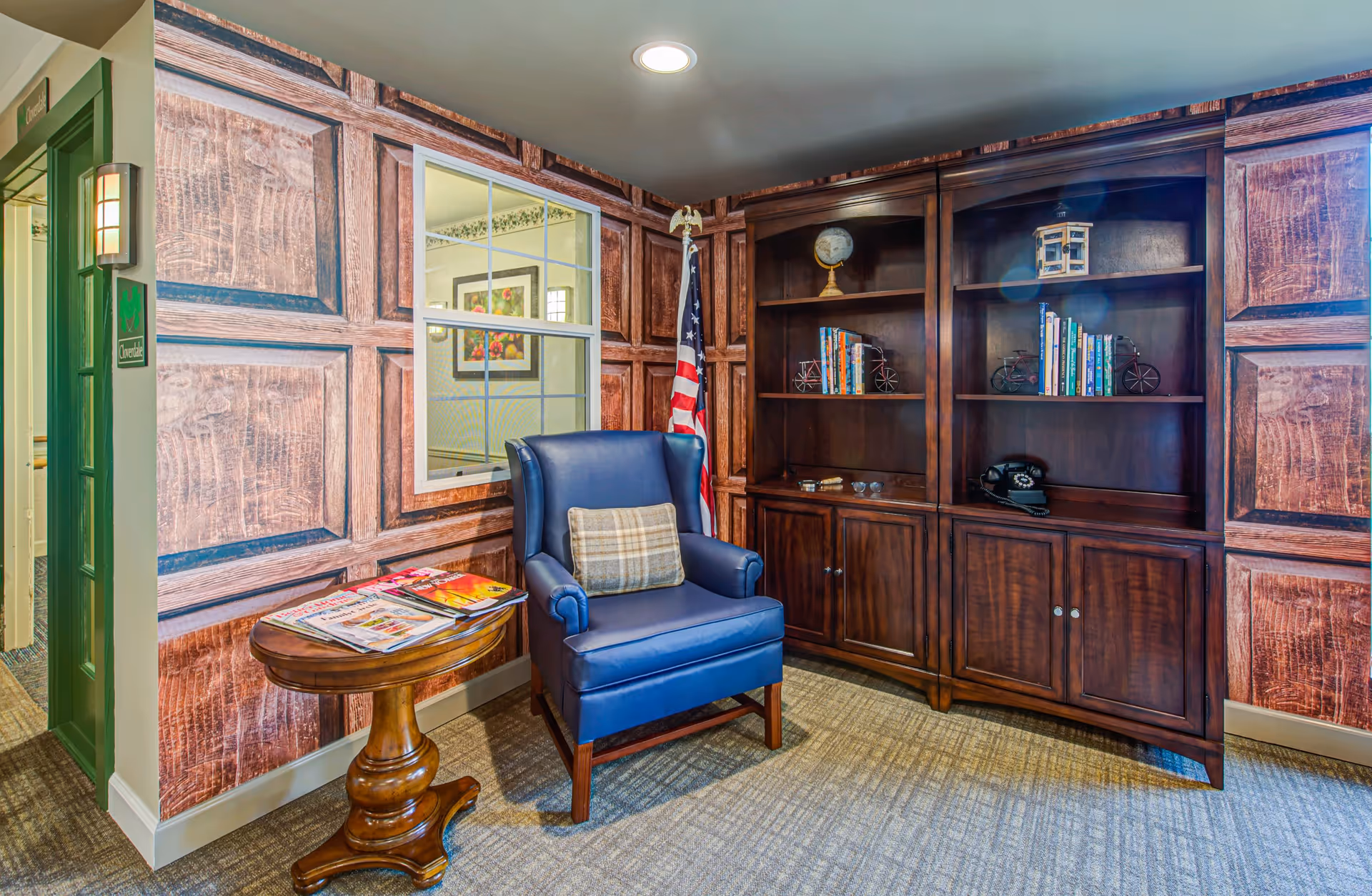 A cozy corner of a senior living community room featuring a blue armchair with a plaid cushion, a small wooden side table with magazines, and a large dark wooden bookshelf with books, decorative items, and a vintage telephone. The walls have a wood panel design, and an American flag stands in the corner. A window looks into another room with framed artwork visible.