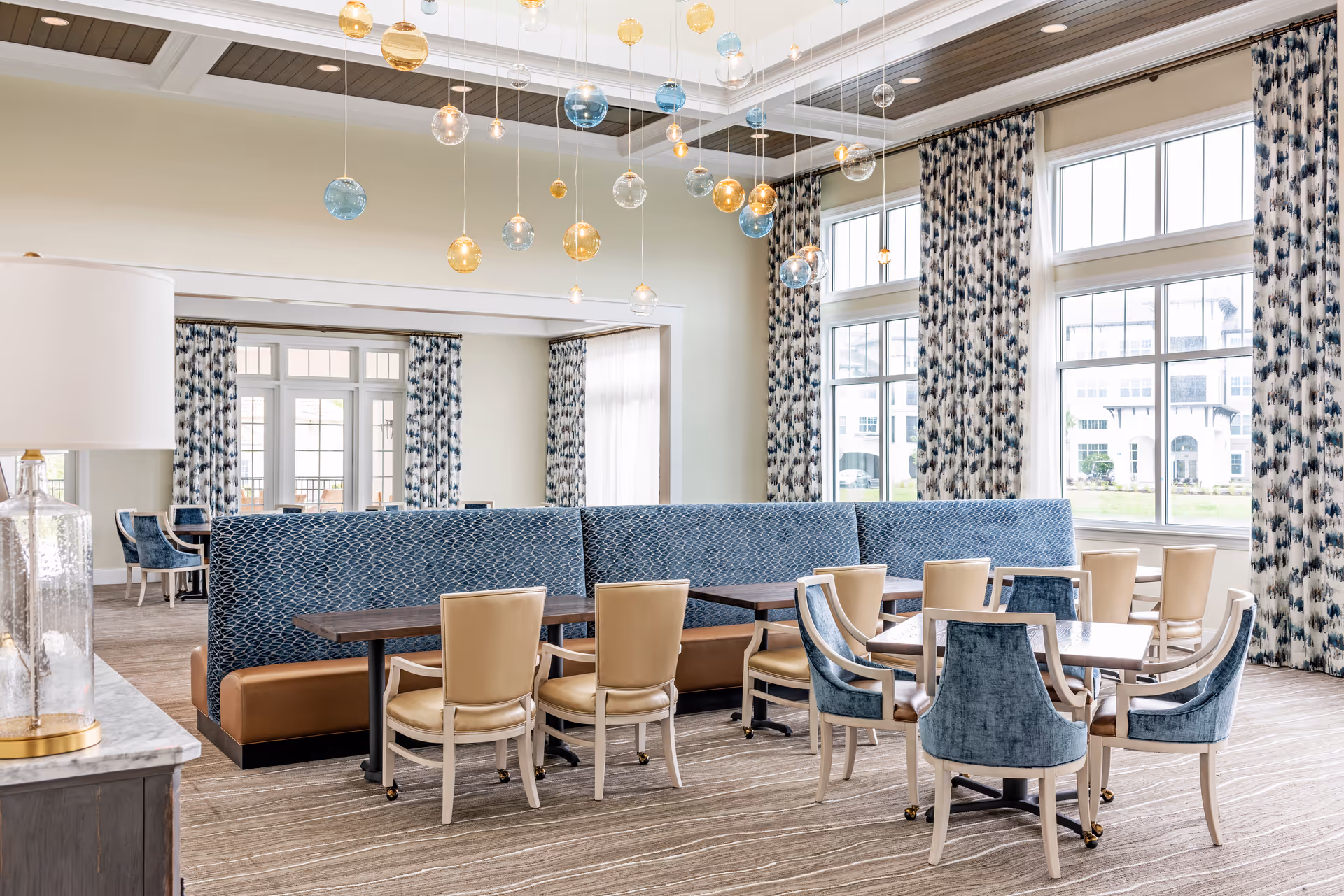 Bright and spacious dining area with large windows covered by patterned curtains, multiple tables with beige and blue upholstered chairs, a long blue cushioned bench, and decorative hanging glass orbs from the ceiling.