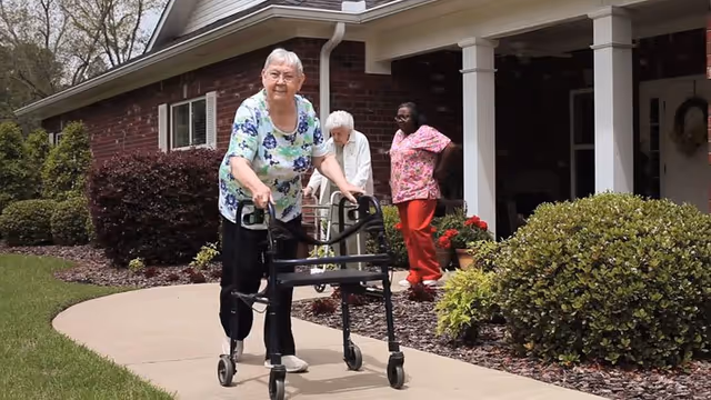 An elderly woman using a walker on a sidewalk outside a brick building with two other women standing nearby, one elderly and one younger, with bushes and plants around the walkway.