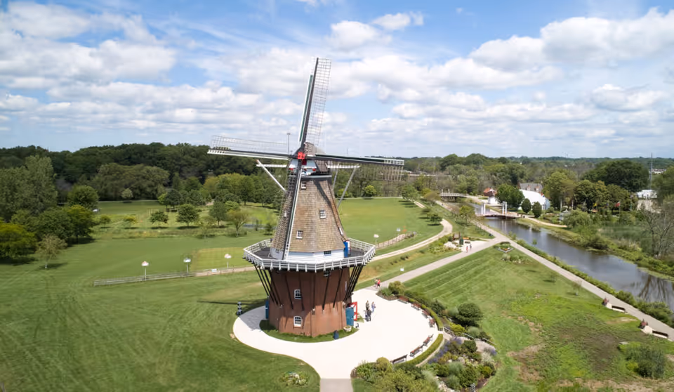 Aerial view of a traditional windmill surrounded by green grass, trees, and walking paths under a partly cloudy sky. A river runs alongside the park area with benches and landscaping.