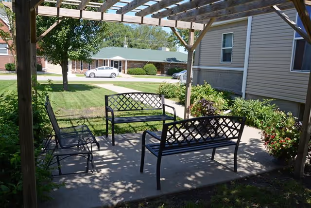 Outdoor seating area under a wooden pergola with three black metal benches arranged on a concrete patio. Surrounding the patio are green bushes and plants, with a lawn and trees visible in the background along with a street and parked cars.