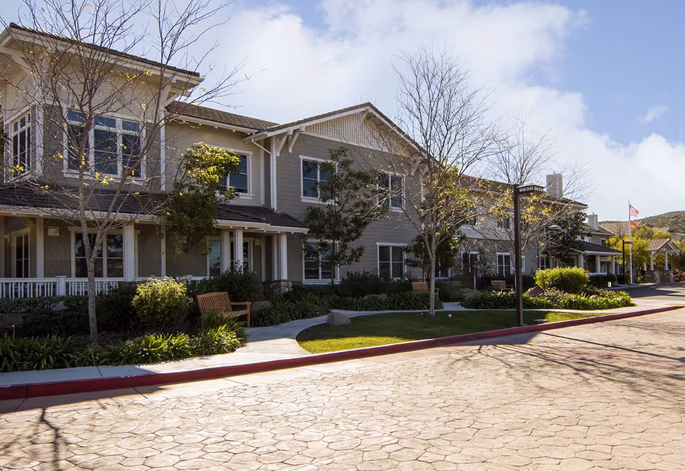 Two-story senior living facility building with landscaped front lawn, benches, walkway and a paved driveway under a partly cloudy sky.