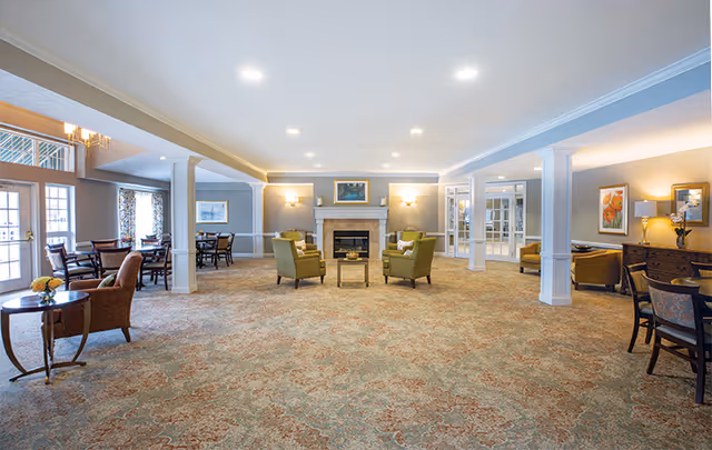 Spacious senior living facility common area with patterned carpet, beige walls, and recessed ceiling lights. The room features a central seating arrangement with four green armchairs around a small table in front of a fireplace. Additional seating areas with tables and chairs are visible on the left and right sides of the room. Decorative paintings and lamps adorn the walls, and large windows allow natural light to enter.