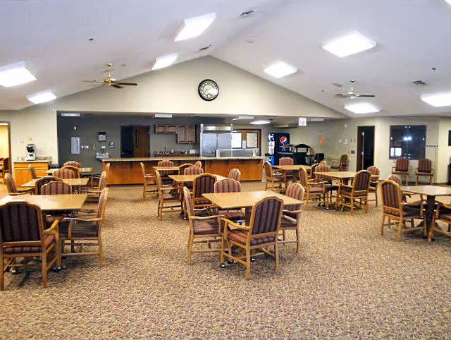 A spacious dining area with multiple wooden tables and chairs arranged neatly on a patterned carpet. The room has a high ceiling with ceiling fans and bright overhead lights. At the far end, there is a kitchen area with stainless steel appliances, a clock on the wall, and a Pepsi vending machine.
