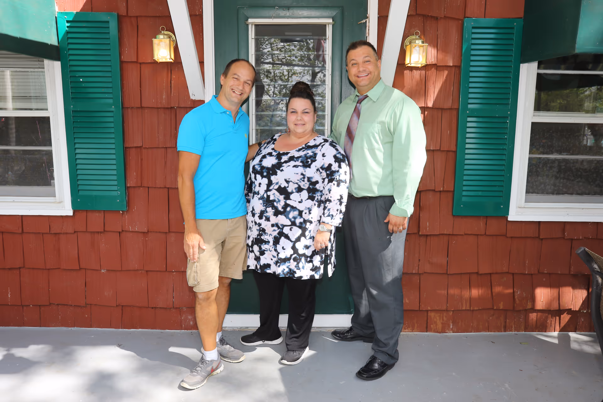 Three adults standing together on a porch in front of a red-shingled building entrance with green shutters.