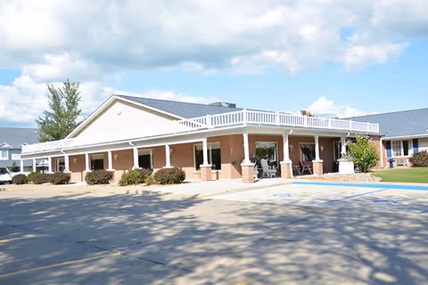 Single-story brick care center building with a covered wraparound porch, entrance seating, and a parking lot under a partly cloudy sky.