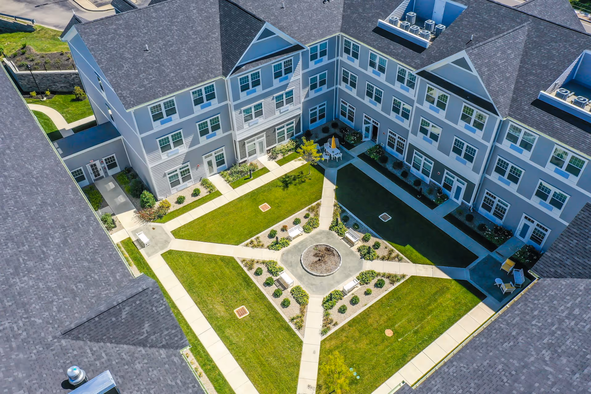 Aerial view of a senior living facility courtyard with well-maintained green lawns, paved walkways, benches, and patio seating areas surrounded by a multi-story building with numerous windows.