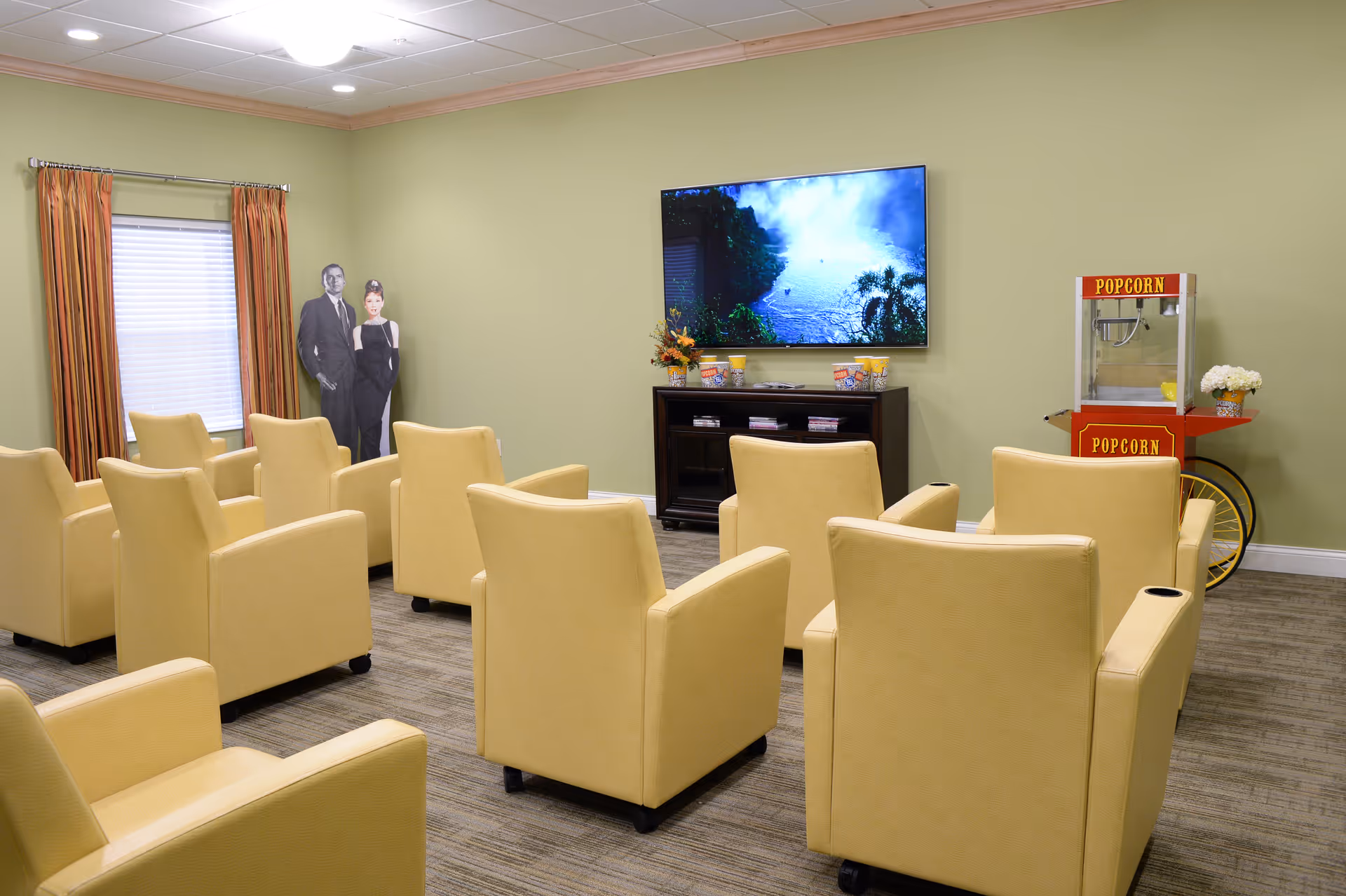 A small theater room with two rows of beige armchairs facing a large flat-screen TV mounted on a light green wall. A popcorn machine is positioned to the right of the TV, and a window with orange curtains is on the left. There is a black and white cardboard cutout of a man and woman standing near the window.