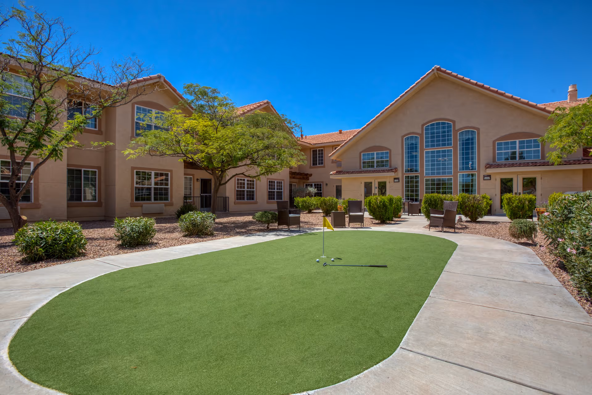Outdoor courtyard area of Vista Pointe at Green Valley featuring a putting green with golf clubs and a flag, surrounded by a paved walkway, shrubs, trees, and beige two-story buildings with large windows under a clear blue sky.