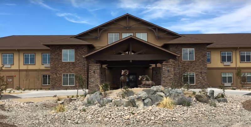 Front exterior view of Volante Senior Living of Reno building with a stone and beige facade, a covered entrance supported by wooden beams, and two ram statues on a rock garden in front. The sky is clear with some clouds.
