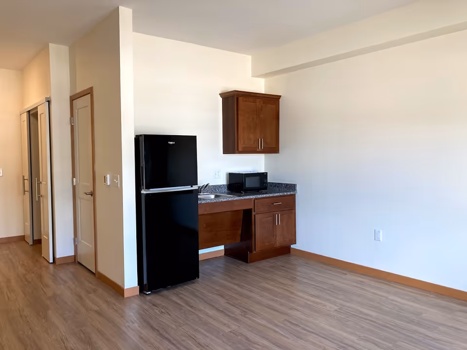 A small kitchenette area in a senior living facility with a black refrigerator, a microwave on a granite countertop, a small sink, and wooden cabinets. The room has light-colored walls and wood flooring, with a hallway and closet doors visible to the left.