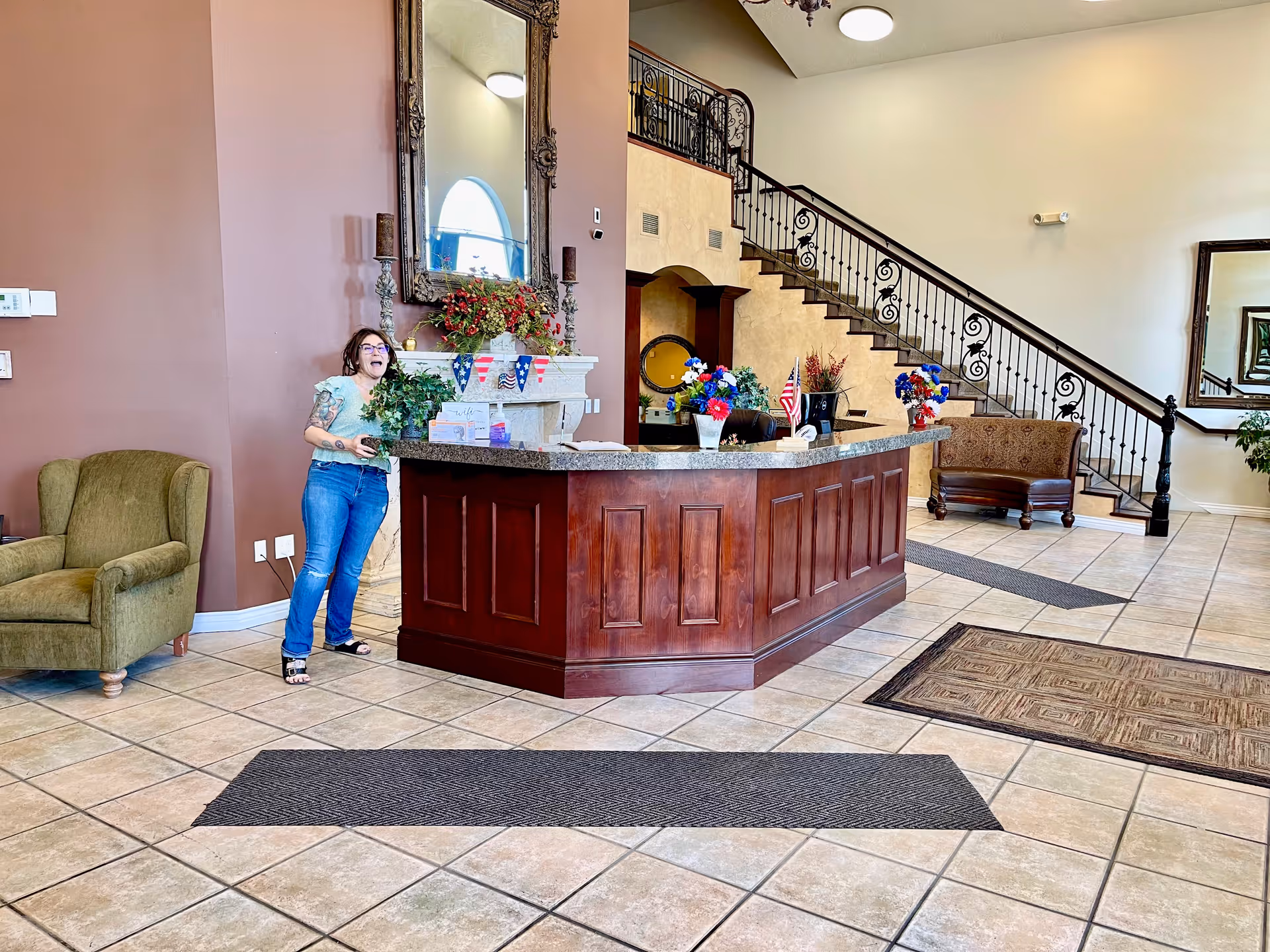 Lobby reception area with a wooden front desk, a staircase, seating, and a person standing to the left holding a plant.