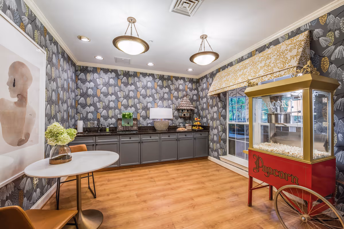 A cozy interior room with patterned wallpaper featuring leaves, a popcorn machine on wheels filled with popcorn, a small round table with two chairs and a vase of flowers, and a long counter with cabinets, a lamp, and decorative items including a birdhouse and a fruit stand.