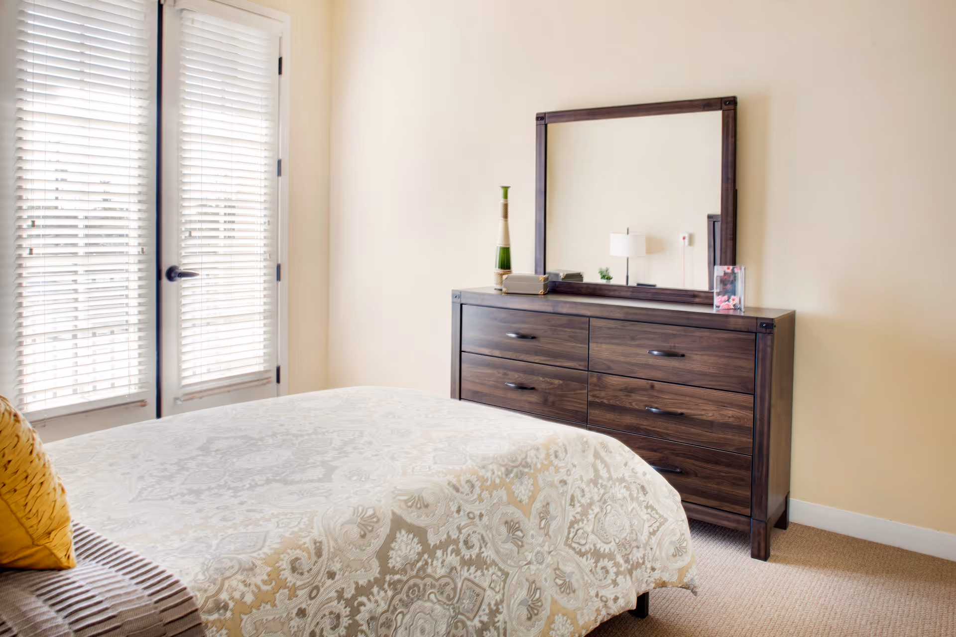 A bright bedroom with a bed covered in a patterned beige and white bedspread, a yellow pillow, and a wooden dresser with a large mirror on top. There is a tall decorative vase and a small framed photo on the dresser. French doors with white blinds allow natural light into the room.