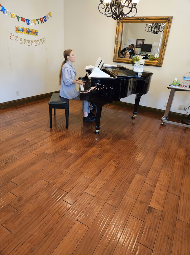 A young woman playing a black grand piano in a room with wooden flooring. There is a large ornate mirror on the wall reflecting the piano and a person in the background. Birthday decorations are hanging on the wall, and a small table with water bottles and other items is visible on the right side.