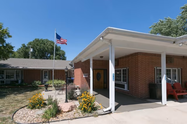 Exterior view of a single-story brick building with a covered entrance supported by white columns. There is a small landscaped area with flowers and rocks near the entrance, and an American flag on a flagpole. The sky is clear and blue.