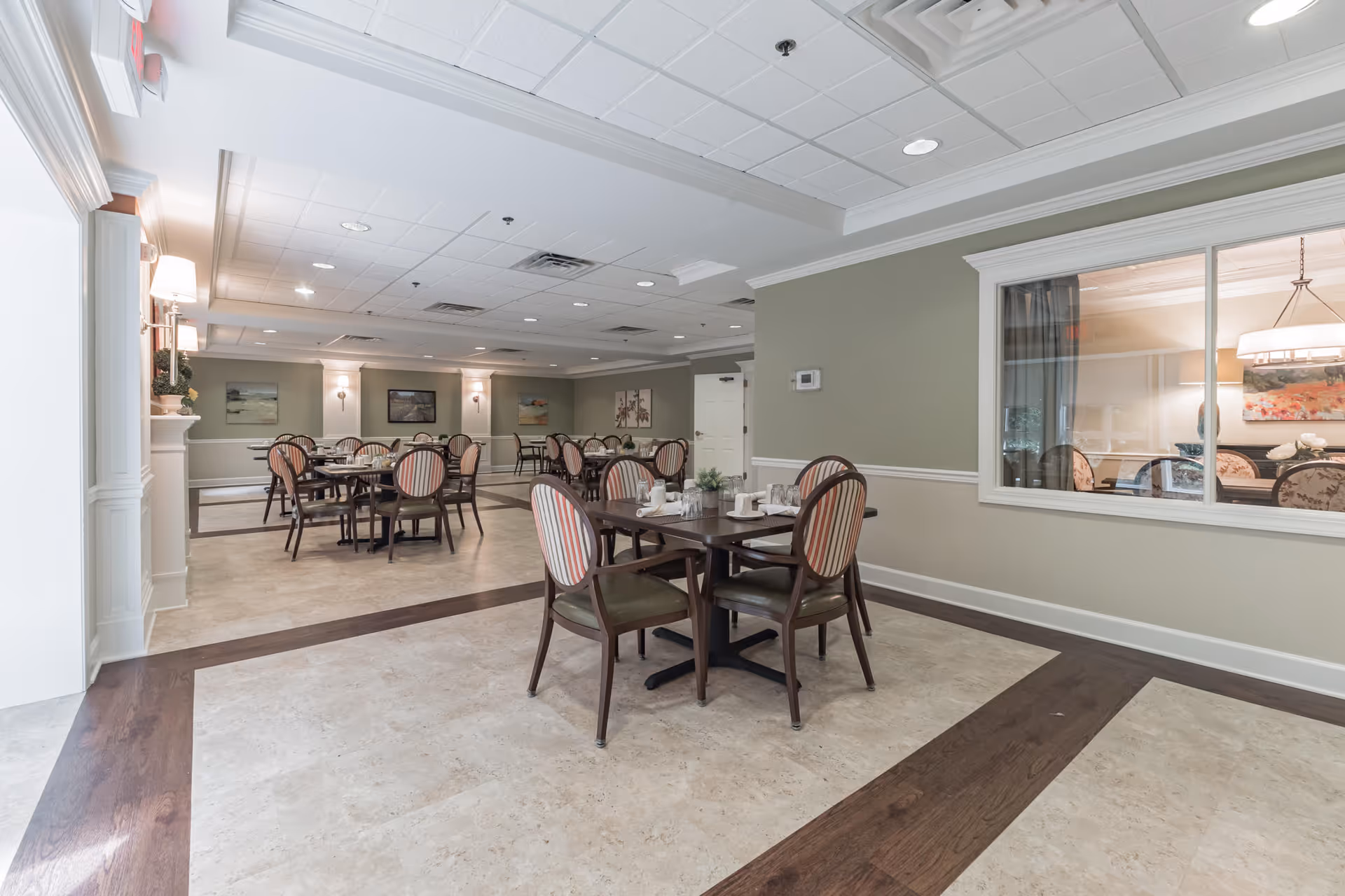 A spacious dining room with multiple tables and chairs arranged neatly. The chairs have striped upholstery and the tables are set with white napkins and small plants. The room features soft green walls with white trim, framed artwork, and a large window looking into another room with similar decor. The ceiling has recessed lighting and a tiled design.