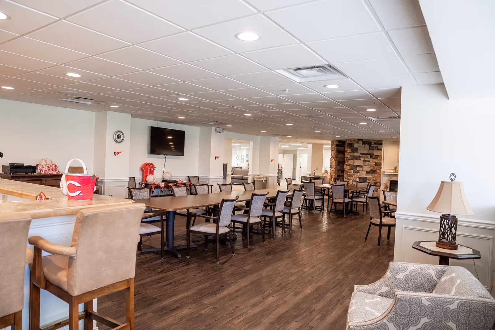 A spacious dining area in a senior living facility featuring multiple tables and chairs arranged neatly on a wooden floor. The room has a white ceiling with recessed lighting, a wall-mounted TV, and a stone accent wall with a fireplace. There are comfortable armchairs and a small table with a lamp in the foreground, and a counter with bar stools on the left side.