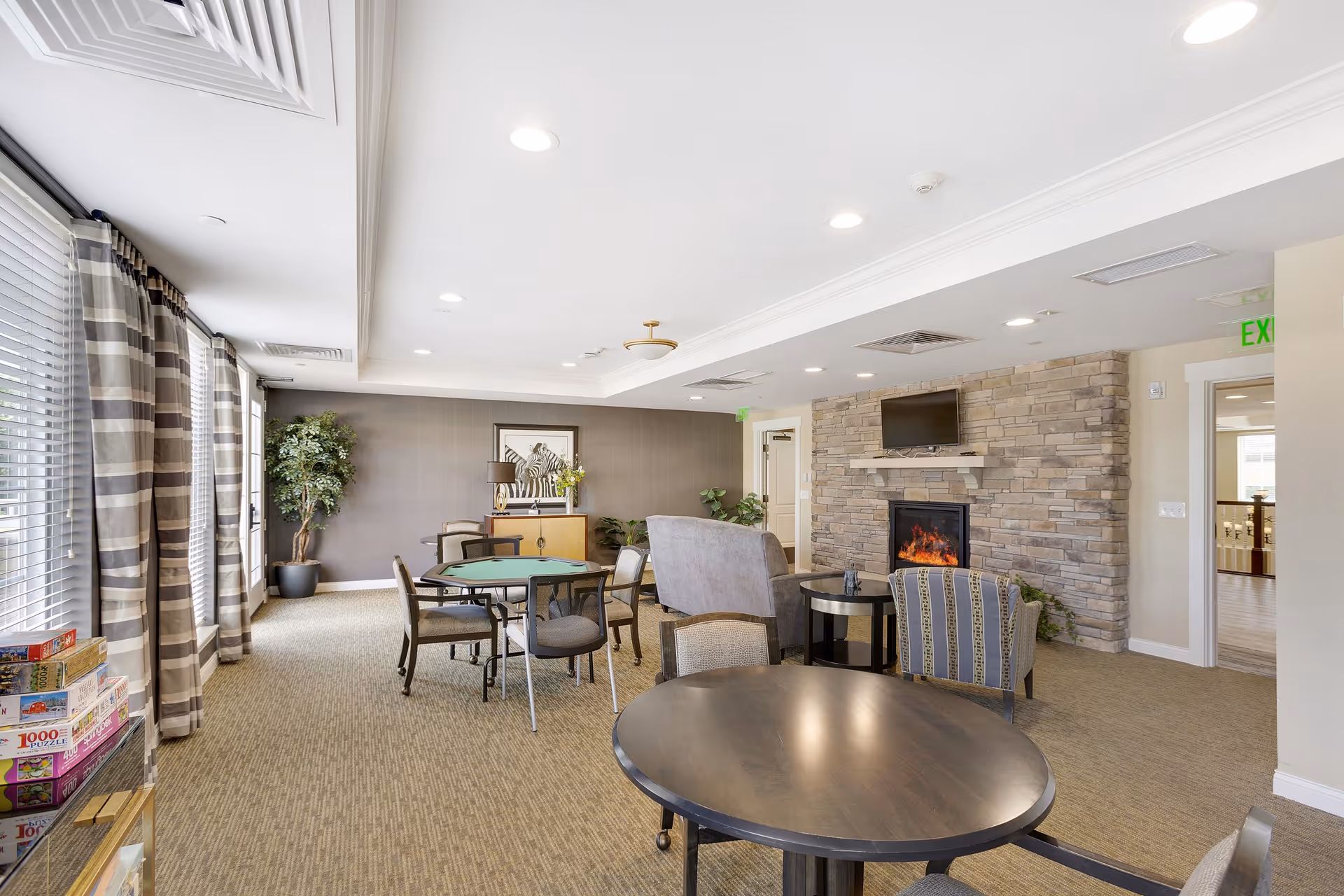 A bright and spacious common area in a senior living facility featuring multiple round tables with chairs, a stone fireplace with a TV mounted above it, large windows with striped curtains, and a few potted plants. Board games and puzzles are stacked on a shelf near the windows.