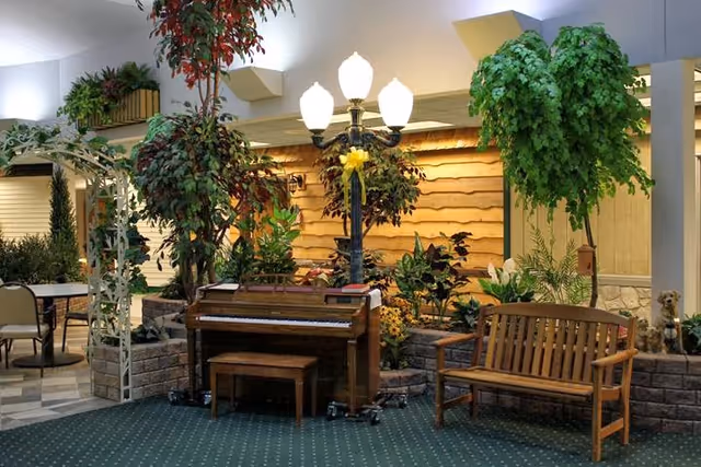 A cozy indoor common area with an upright piano, wooden bench, lamppost, and many potted plants.