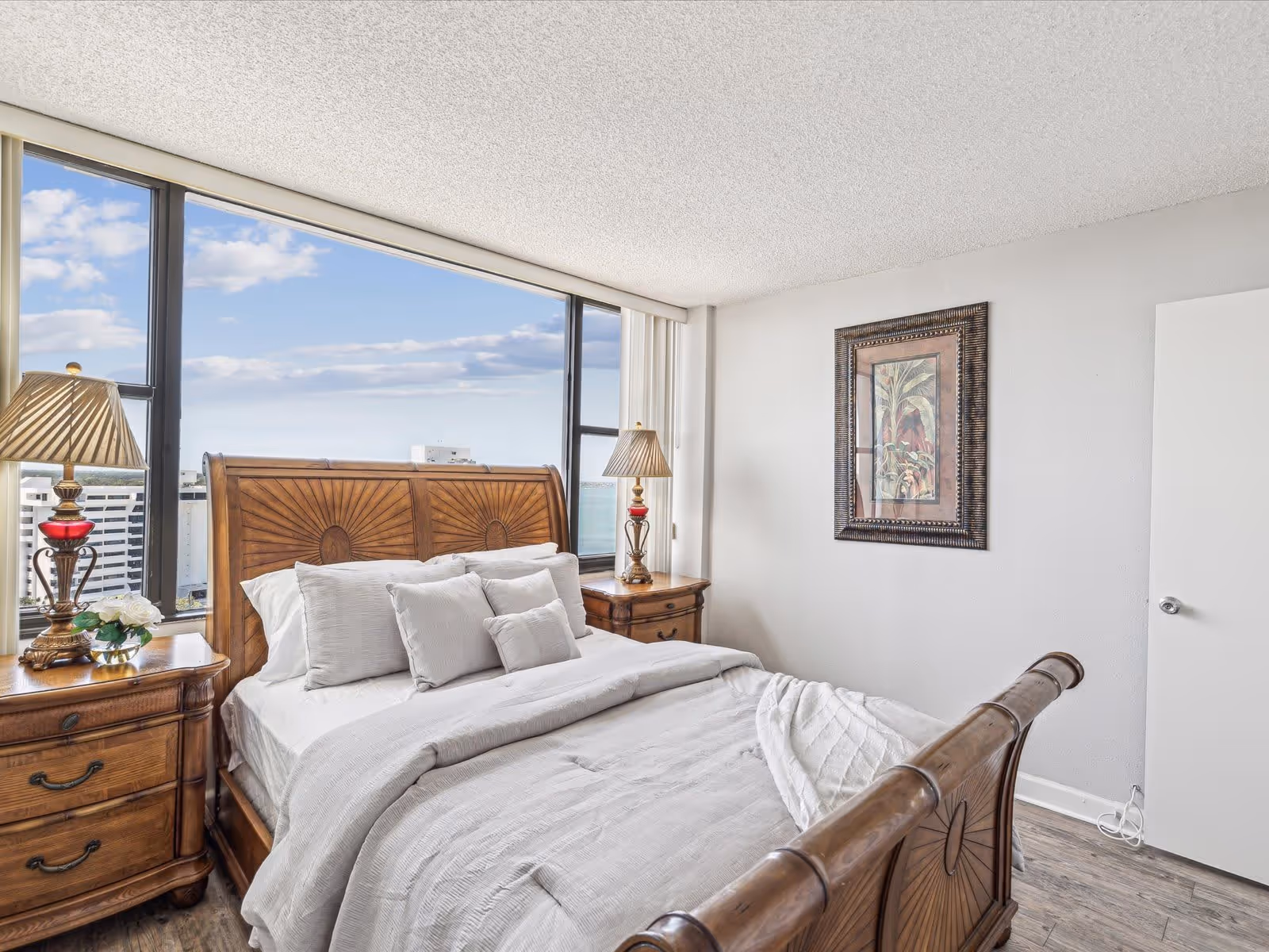 A bright bedroom with a large wooden bed featuring white bedding and multiple pillows. There are two wooden nightstands on either side of the bed, each with a decorative lamp. A framed artwork hangs on the white wall to the right. Large windows behind the bed offer a view of buildings and a partly cloudy sky outside.