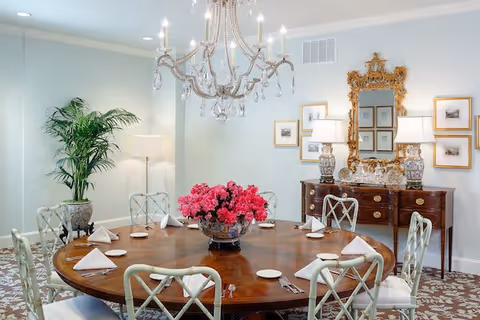 A well-lit dining room with a large round wooden table set with white napkins, plates, and silverware. The room features a crystal chandelier hanging above the table, a decorative sideboard with two ornate lamps and a large gold-framed mirror, framed pictures on the wall, a potted plant in the corner, and a patterned carpet.