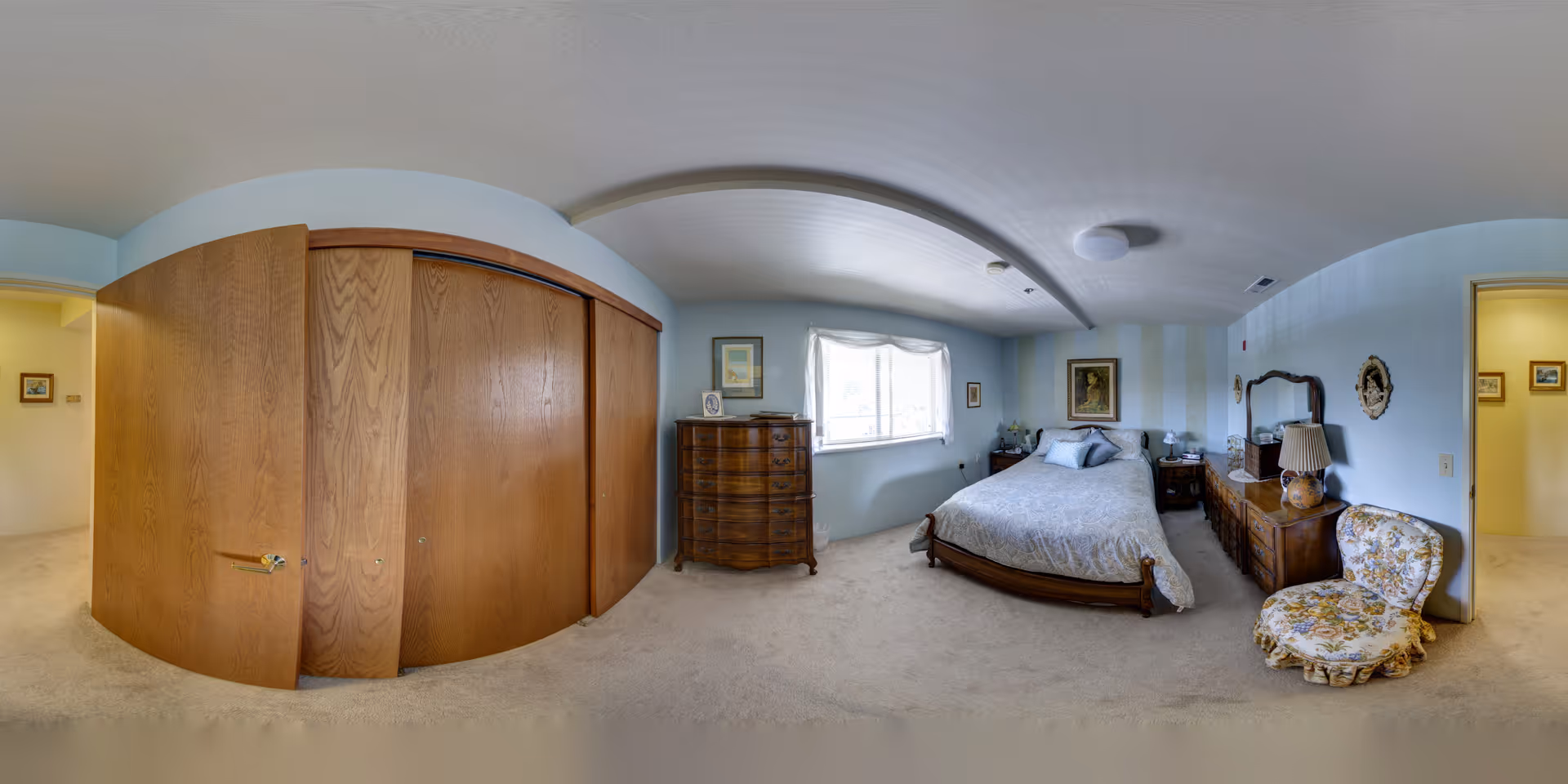 Spacious bedroom with a bed, wooden dressers, a large built-in closet, a window, and a floral armchair.