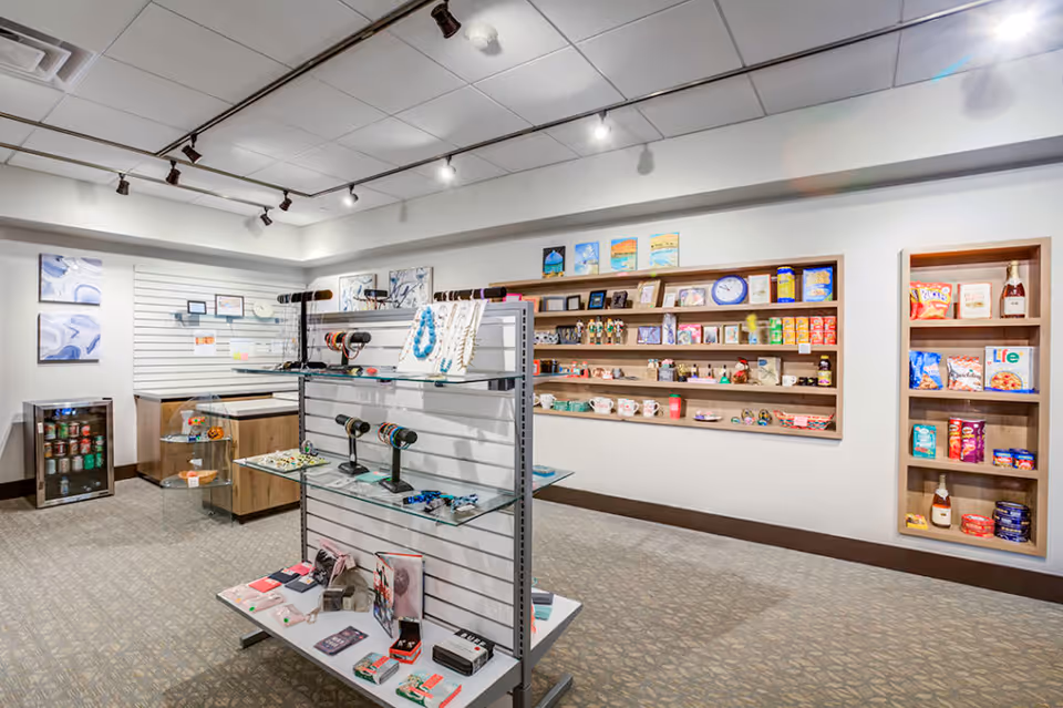Interior view of a small retail shop area inside a senior living facility named The Palazzo, featuring shelves and display racks with various items including jewelry, snacks, canned goods, and small gift items. The room has a carpeted floor, white walls, and ceiling track lighting.