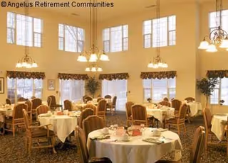 Spacious, high-ceilinged dining room with round tables set for meals, upholstered chairs, chandeliers, and large windows.