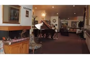 Interior view of a senior living facility lounge area featuring a grand piano, a fireplace with a wooden mantel, a patterned chair, and various framed artworks on the walls. The space is carpeted and well-lit with ceiling lights and a chandelier, with a hallway leading to other rooms in the background.