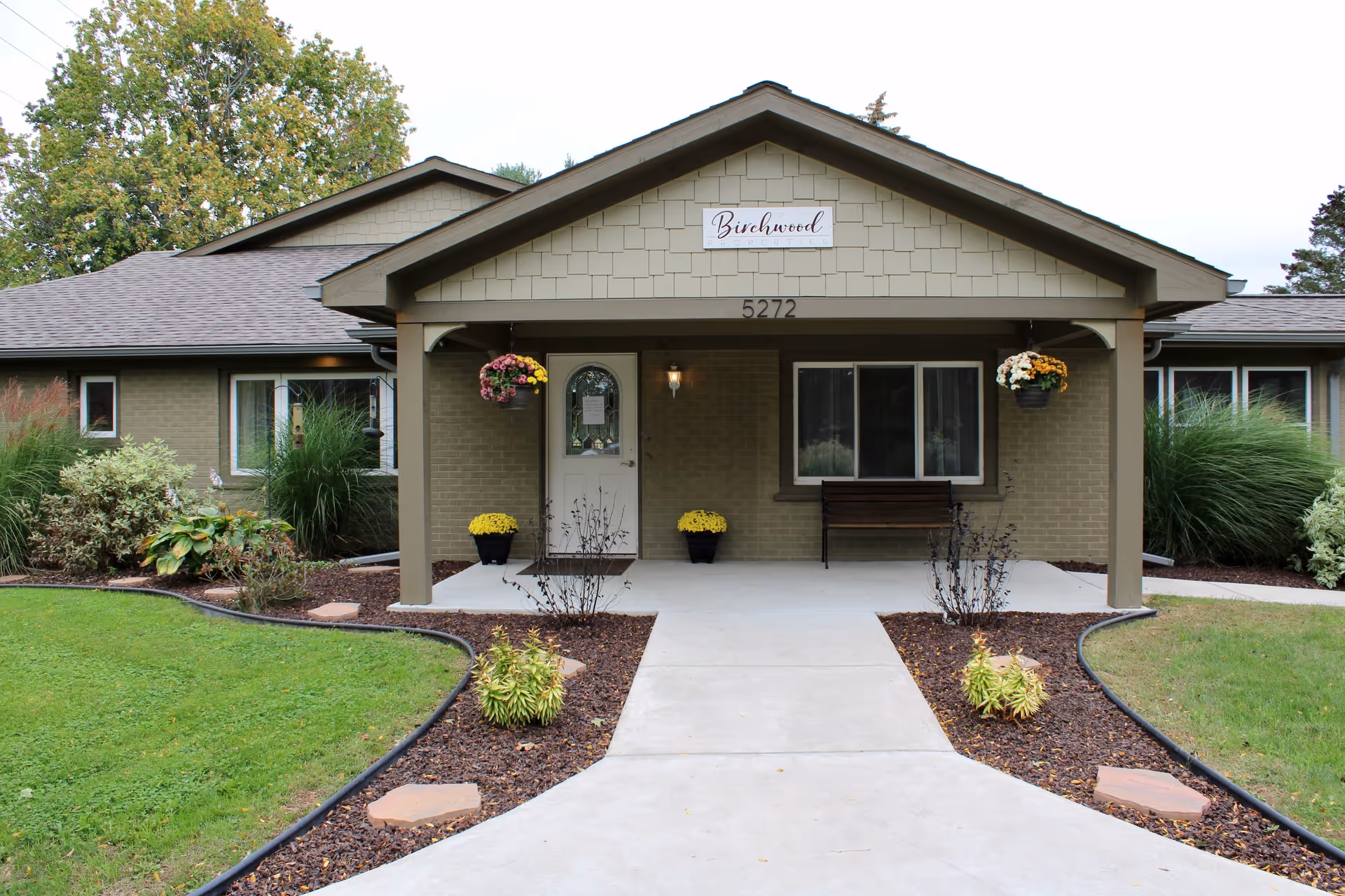 Front exterior view of a single-story building with a covered entrance. The building has a sign above the door that reads 'Birchwood Properties' and the number 5272. There are hanging flower baskets and potted plants near the entrance, a bench to the right of the door, and landscaped garden beds with mulch and small plants along the walkway leading to the door.
