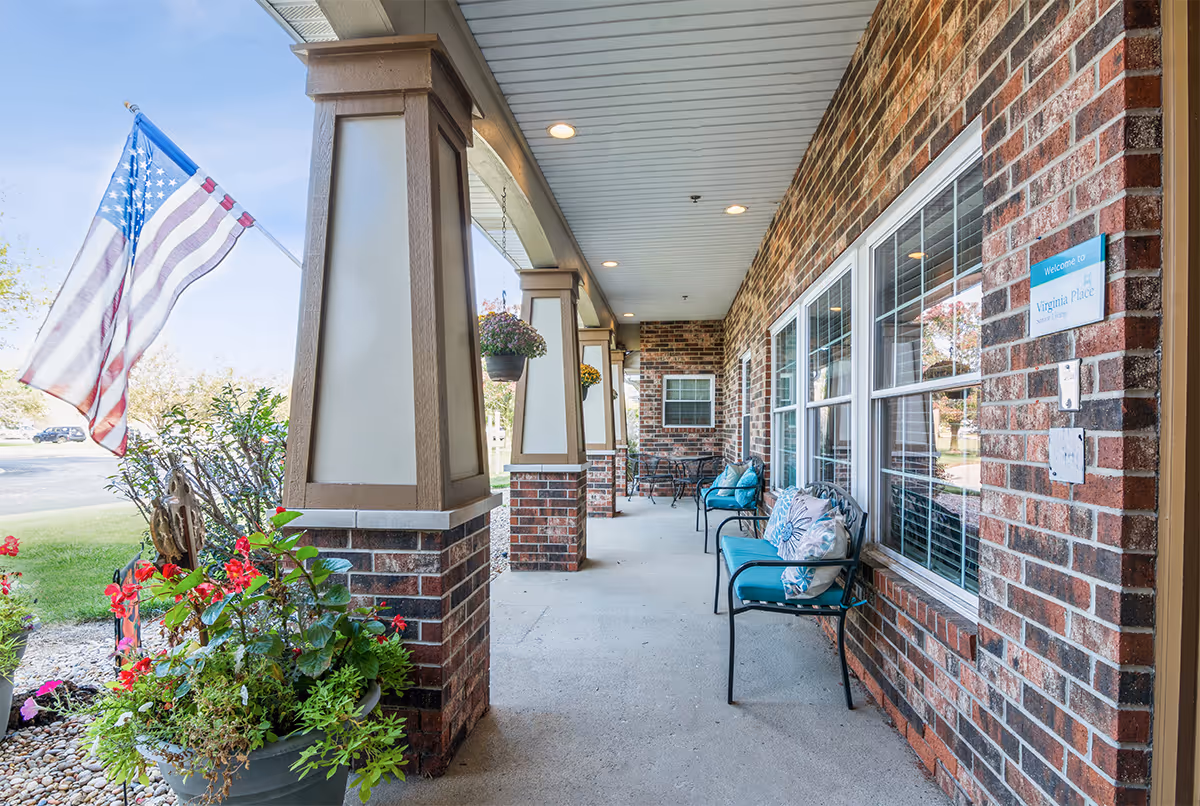 Front covered porch of Virginia Place with brick columns, seating, potted plants, and an American flag.
