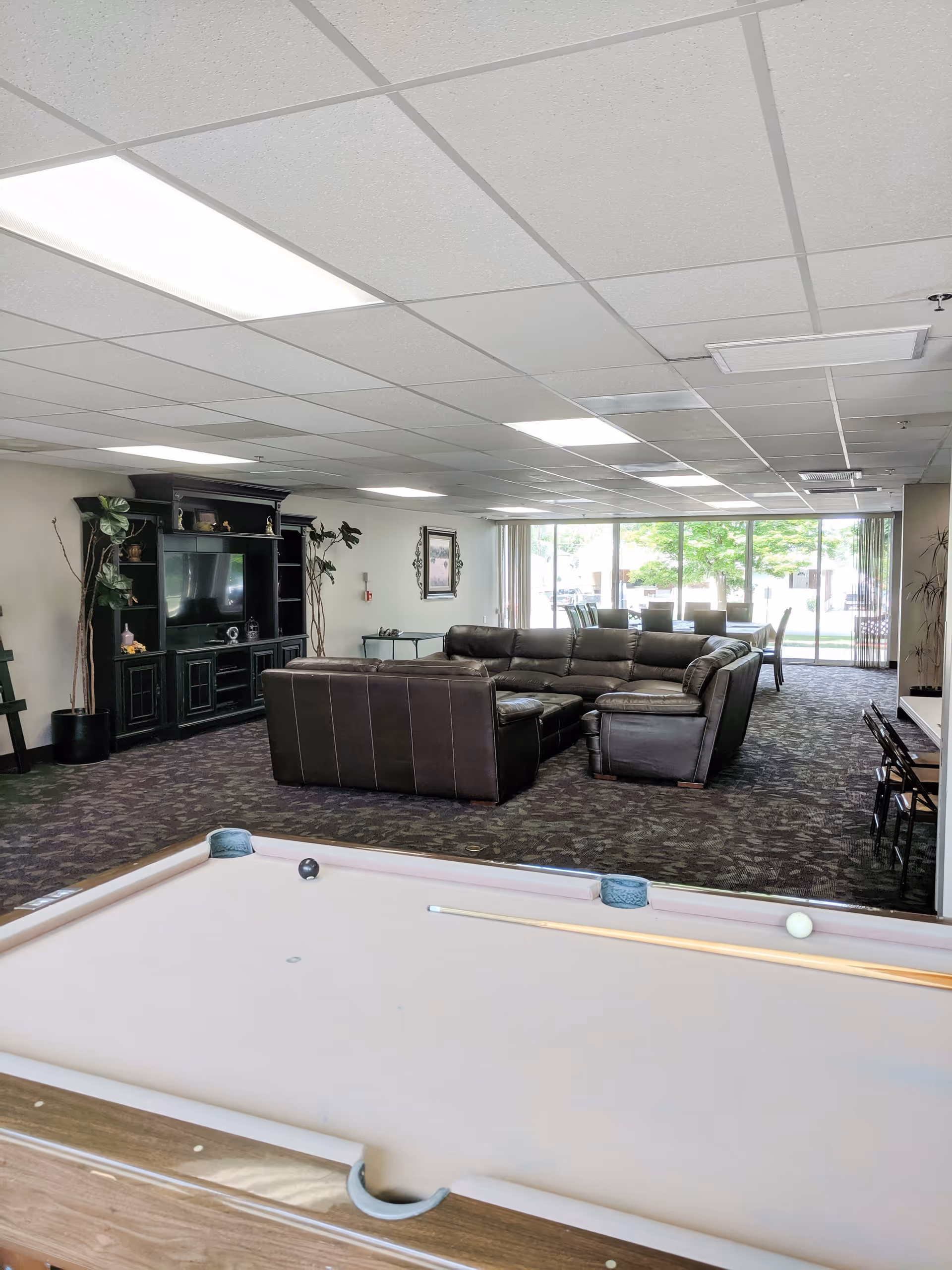 Community lounge with a pool table in the foreground, leather sectional seating, TV cabinet, and dining area by large windows.