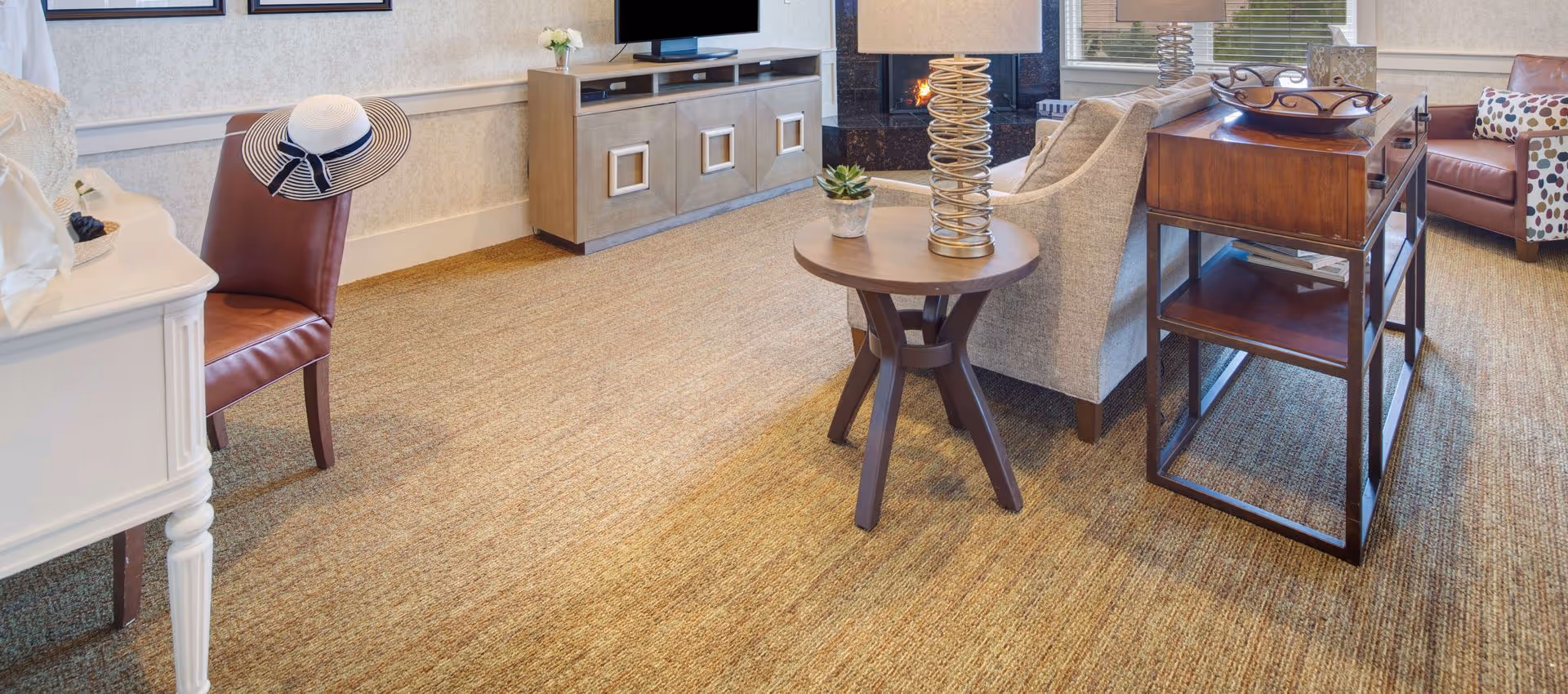 A cozy living room area with a beige carpet, a gray sofa, a wooden side table with a lamp and a small plant, a wooden console table with a decorative tray, a leather chair with a striped hat on it, a fireplace, and large windows with blinds.