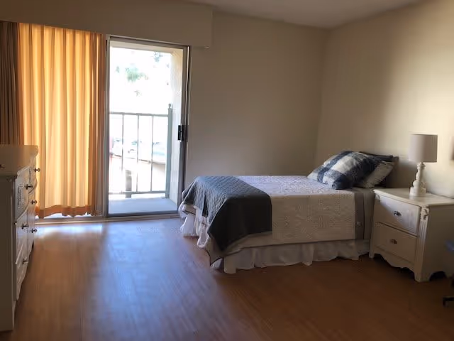 A simple bedroom with a single bed covered with a white quilt and a dark gray throw blanket, a checkered pillow, a white nightstand with a lamp, a dresser, and a sliding glass door with beige curtains leading to a small balcony.