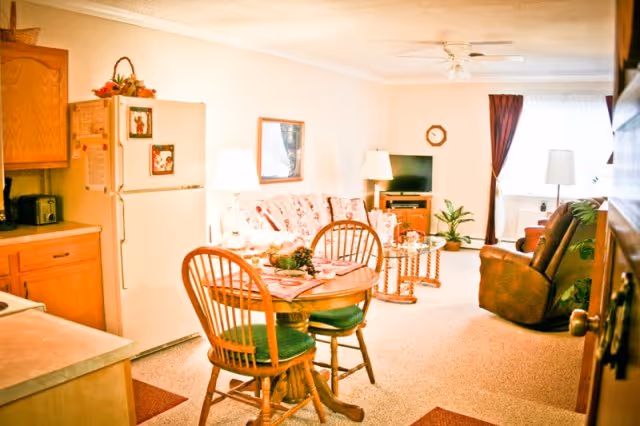 Open-plan living area with a small round dining table and chairs beside a kitchenette, a sofa, TV and recliner in a warmly lit room.