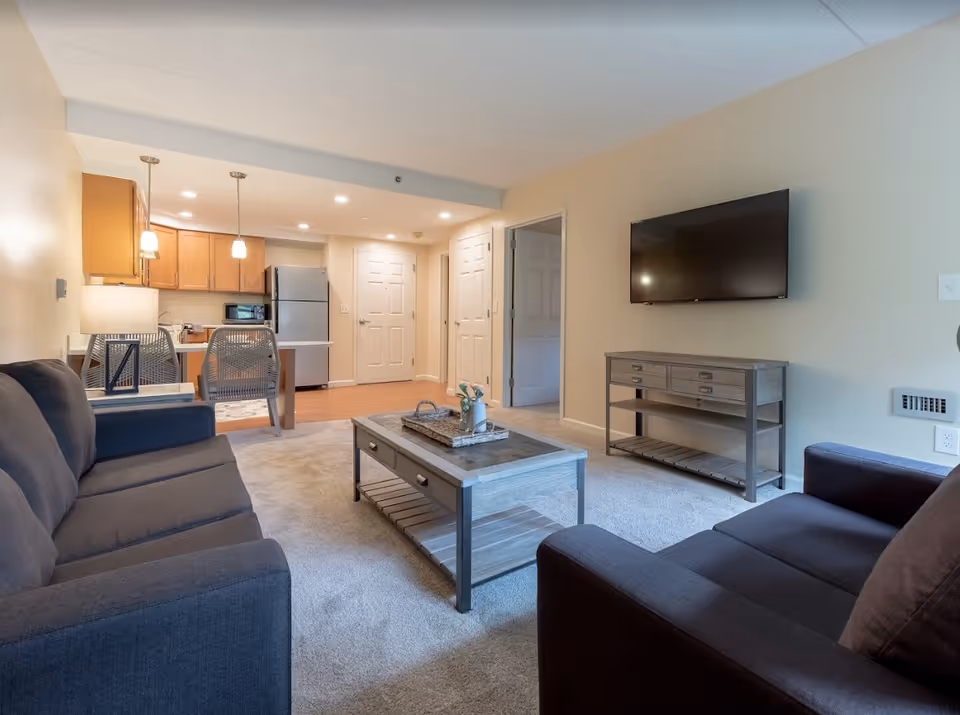 A cozy living room area with two dark sofas facing a wooden coffee table. A flat-screen TV is mounted on the wall above a wooden console table. In the background, there is a kitchen area with wooden cabinets, a refrigerator, a microwave, and two pendant lights hanging over a small dining table with two chairs. The room has beige walls and carpeted flooring in the living area, with hardwood flooring in the kitchen.
