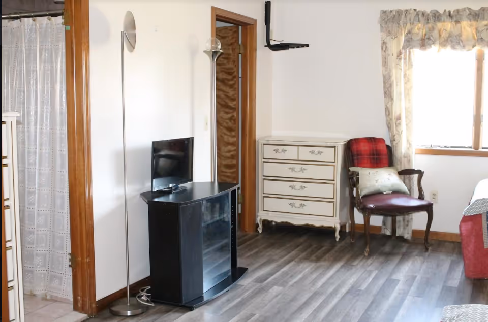 Bright bedroom corner with a small TV on a black stand, white dresser, wooden chair with a blanket, floor lamp, and a partial bed by a window.