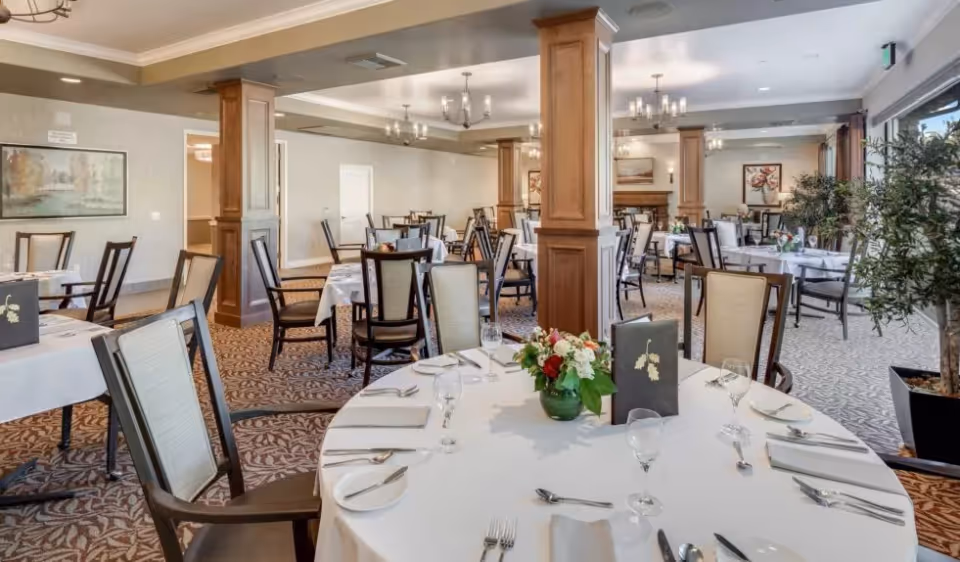 A spacious dining room with multiple round and rectangular tables covered with white tablecloths, set with plates, silverware, napkins, and glasses. The room features wooden columns, patterned carpet, chandeliers, framed artwork on the walls, and large windows with plants near the entrance.