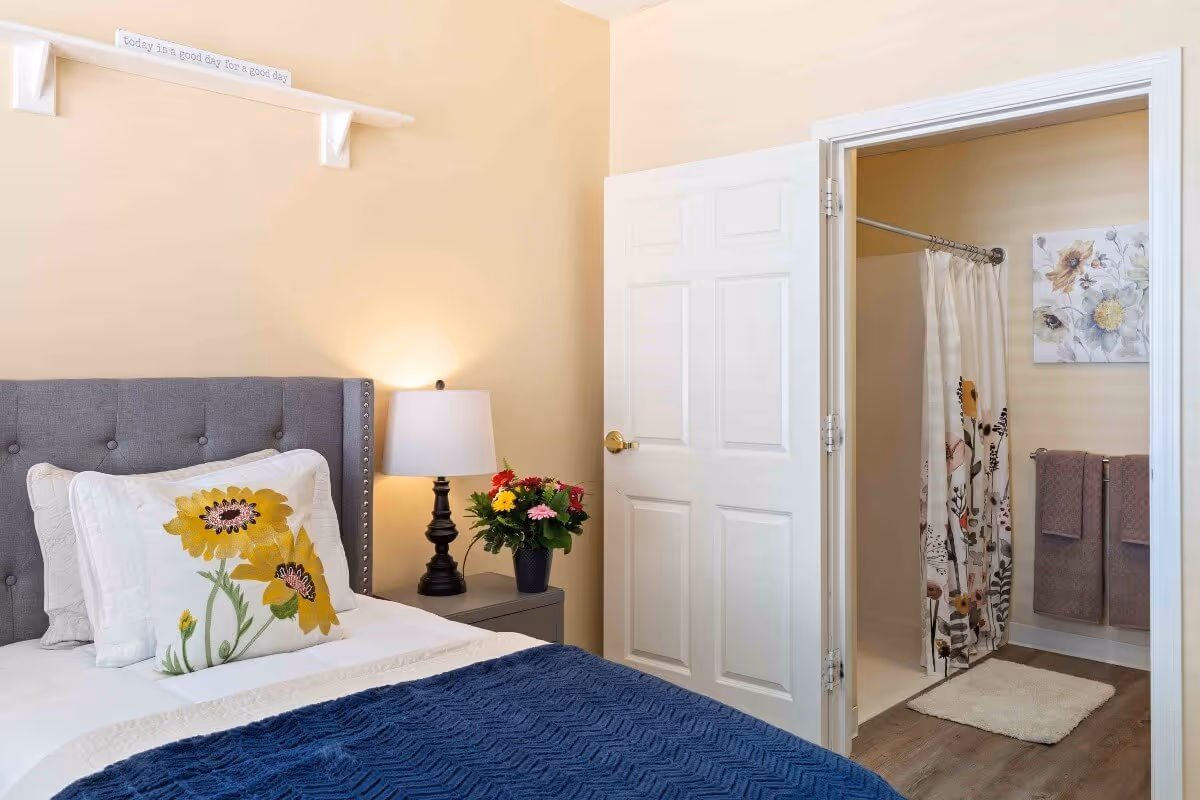 A cozy bedroom with a gray upholstered headboard, white bedding, and a decorative pillow featuring yellow flowers. A nightstand with a black lamp and a vase of colorful flowers is next to the bed. The door to an adjoining bathroom is open, showing a shower with a floral curtain, two brown towels hanging on a rack, a white bath mat, and a floral wall art. A small shelf above the bed holds a sign that reads 'today is a good day for a good day.'