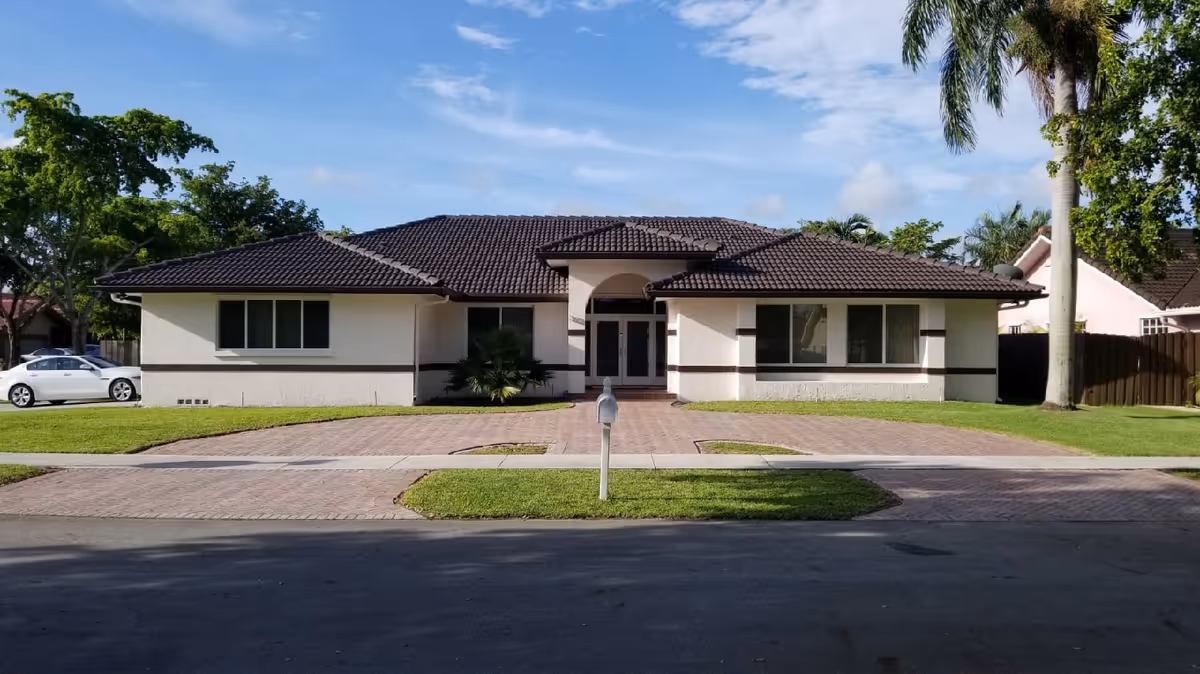 Single-story ranch-style house with a tiled roof, central double-door entrance, brick driveway and mailbox, set on a lawn with palm trees.