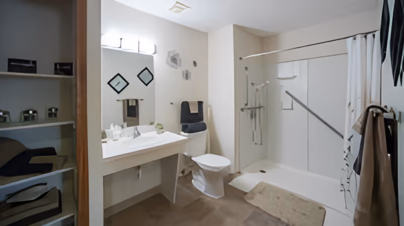 A clean and modern bathroom featuring a white toilet, a large mirror above a white sink with a countertop, a walk-in shower with a grab bar and a shower curtain, beige floor mats, and neatly folded towels on the toilet and hanging on the wall.