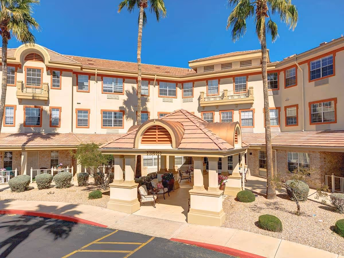 Exterior view of a three-story assisted living facility building with beige walls and red trim around the windows. There is a covered entrance with a tiled roof supported by columns, and several chairs and potted plants underneath. Palm trees and desert landscaping with bushes and gravel surround the building. The sky is clear and blue.