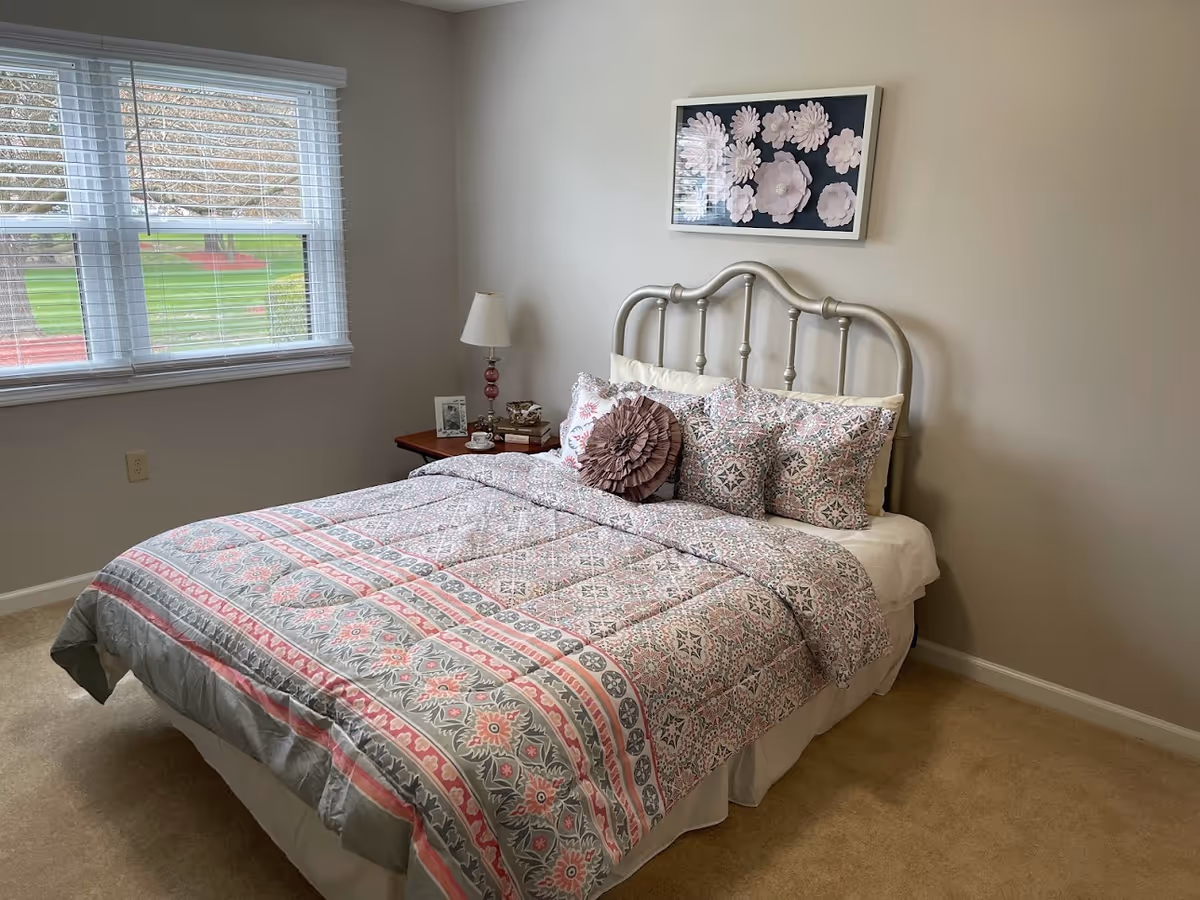 A neatly made bed with a patterned quilt and multiple pillows in a bedroom. The bed has a metal headboard and is positioned against a beige wall with a framed floral artwork above it. A wooden nightstand with a lamp, a framed photo, and some decorative items is next to the bed. A window with white blinds shows a green lawn outside.