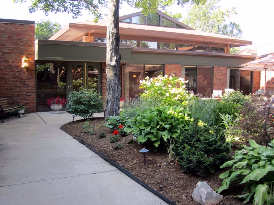 Brick building front with large windows, a paved walkway and a landscaped garden with trees and shrubs.